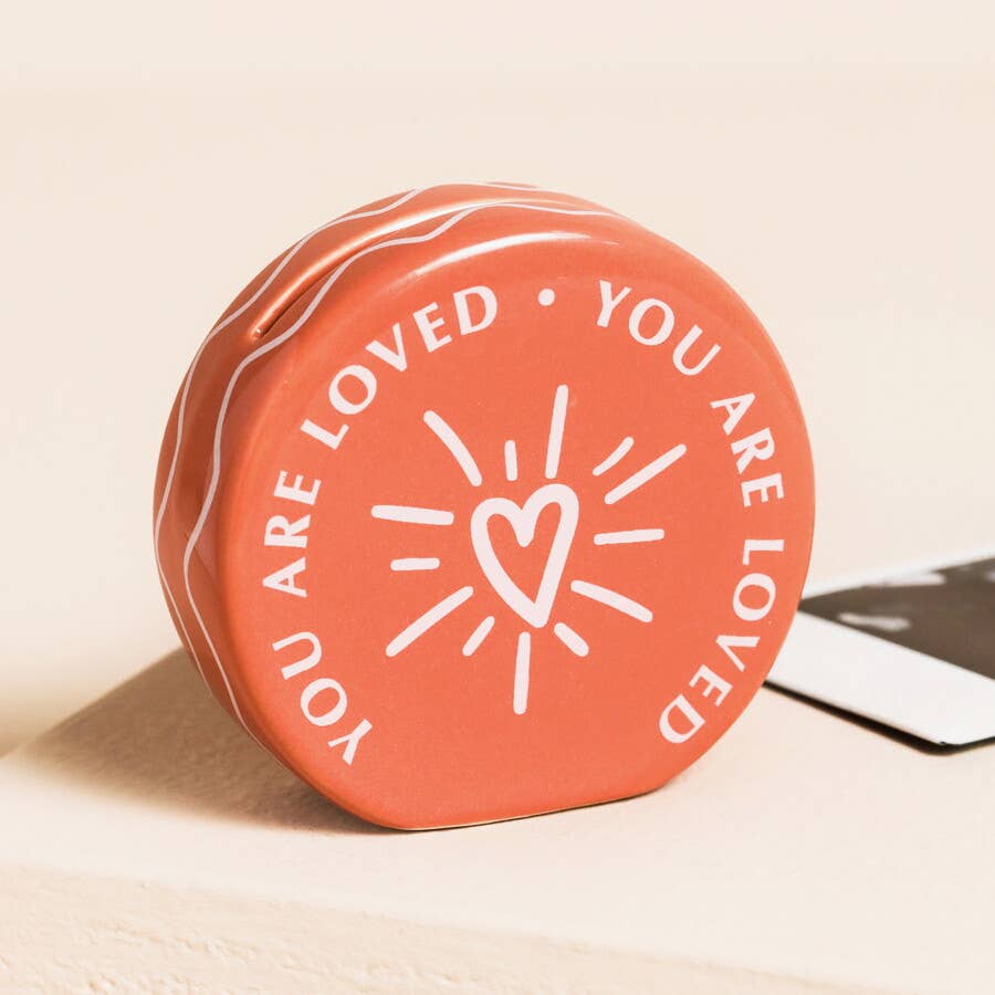An orange ceramic tin with white text reading 'YOU ARE LOVED • YOU ARE LOVED' and a white heart design in the center, photographed on a light beige surface with a blurred card in the background.