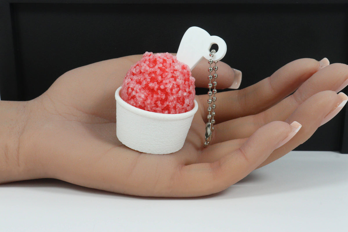 A hand holding a miniature red shaved ice keychain in a white cup with a silver ball chain and white heart-shaped tag.