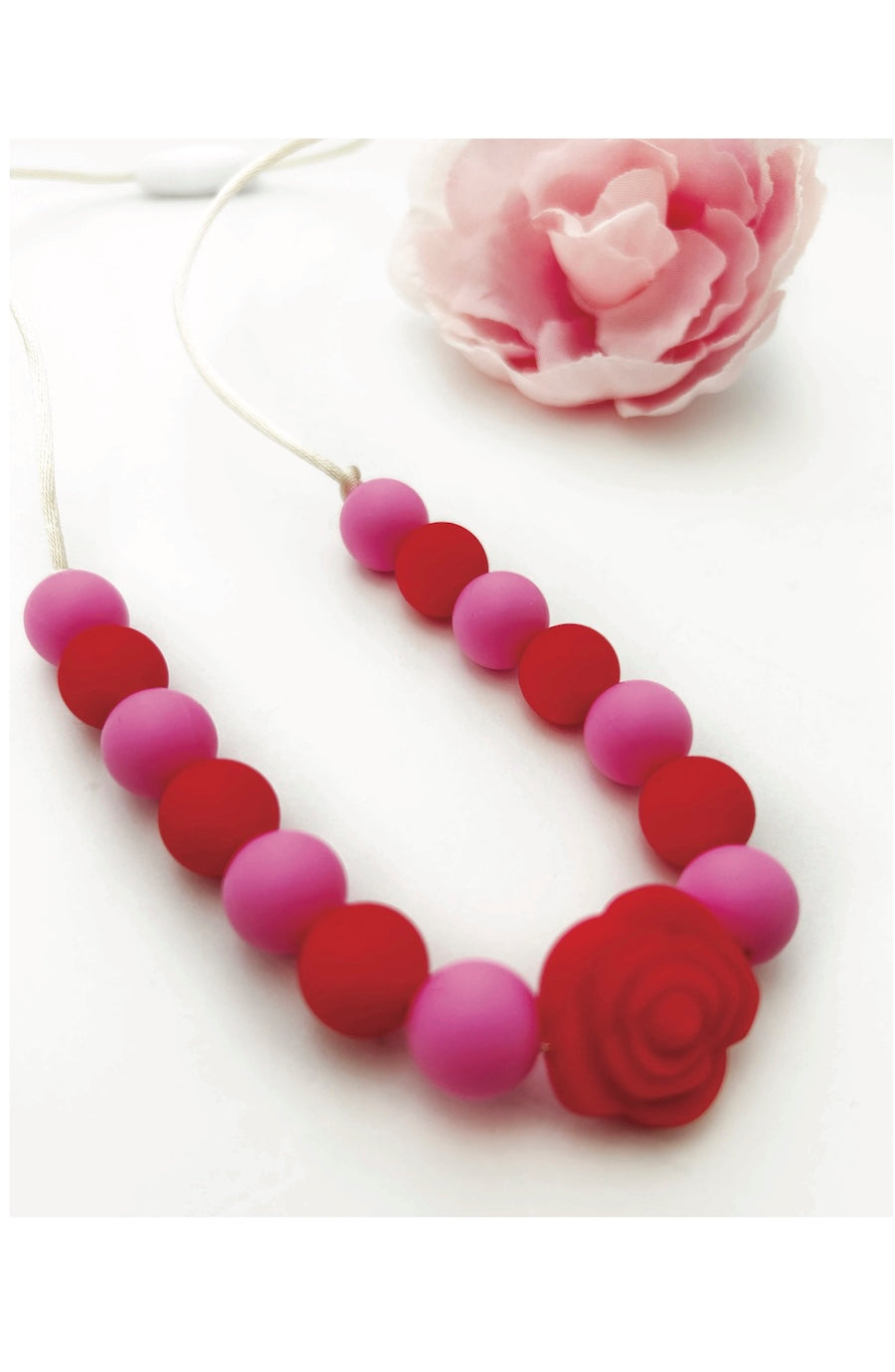 A silicone teething necklace with alternating red and pink round beads, featuring a large red rose charm at one end, displayed on a white background with a decorative pink flower.