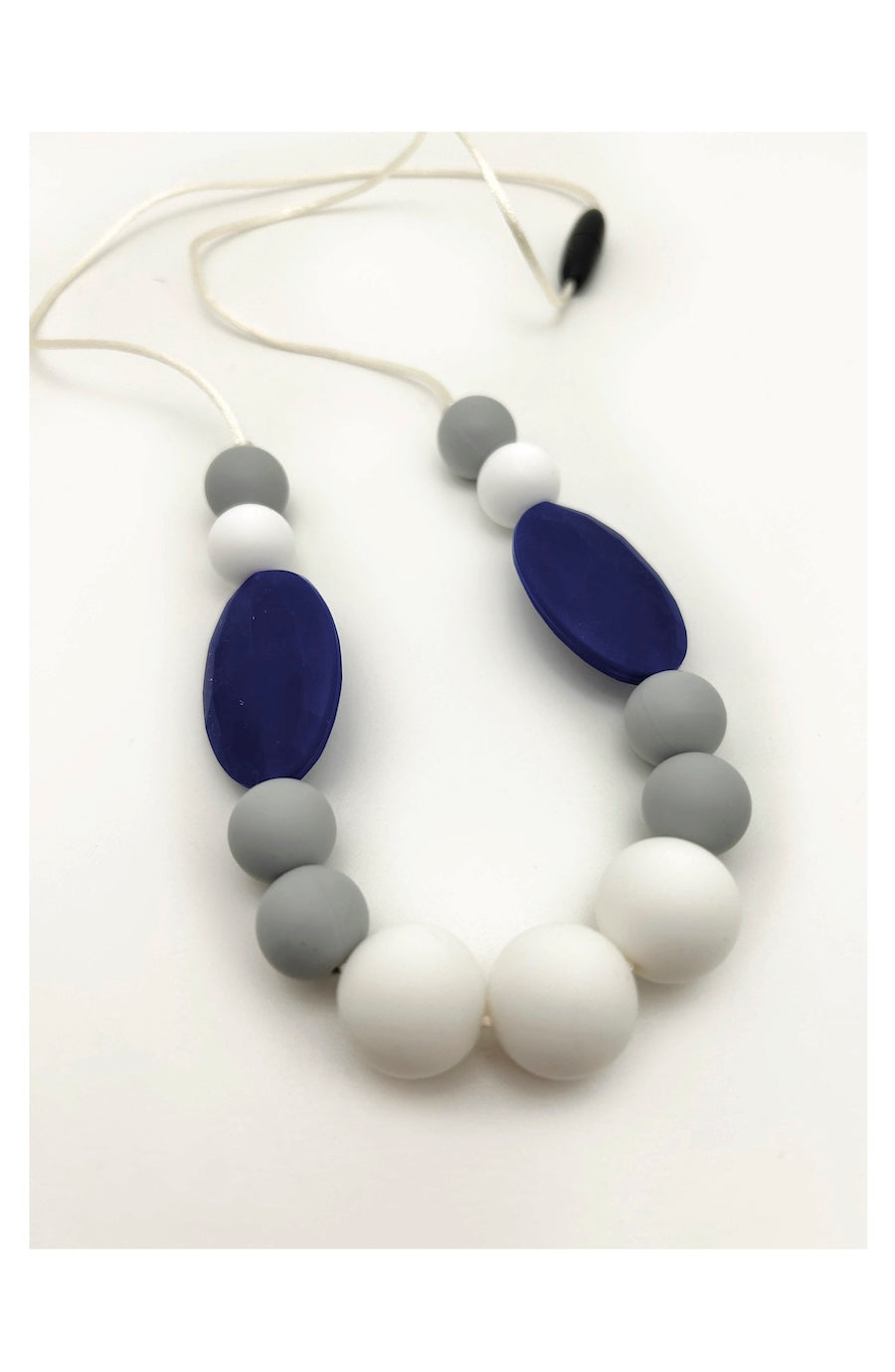 A silicone beaded necklace for babies or toddlers, featuring matte white and light gray round beads, two larger dark blue oval beads, and a white cord with a black toggle clasp, shown on a plain white background.