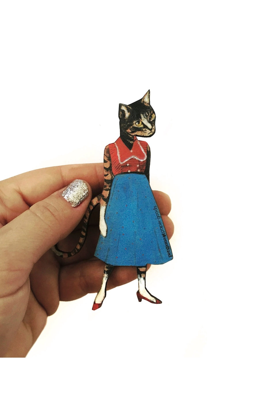 A hand with glittery silver nail polish holds a small illustrated cat figurine wearing a red top and blue skirt against a plain white background.