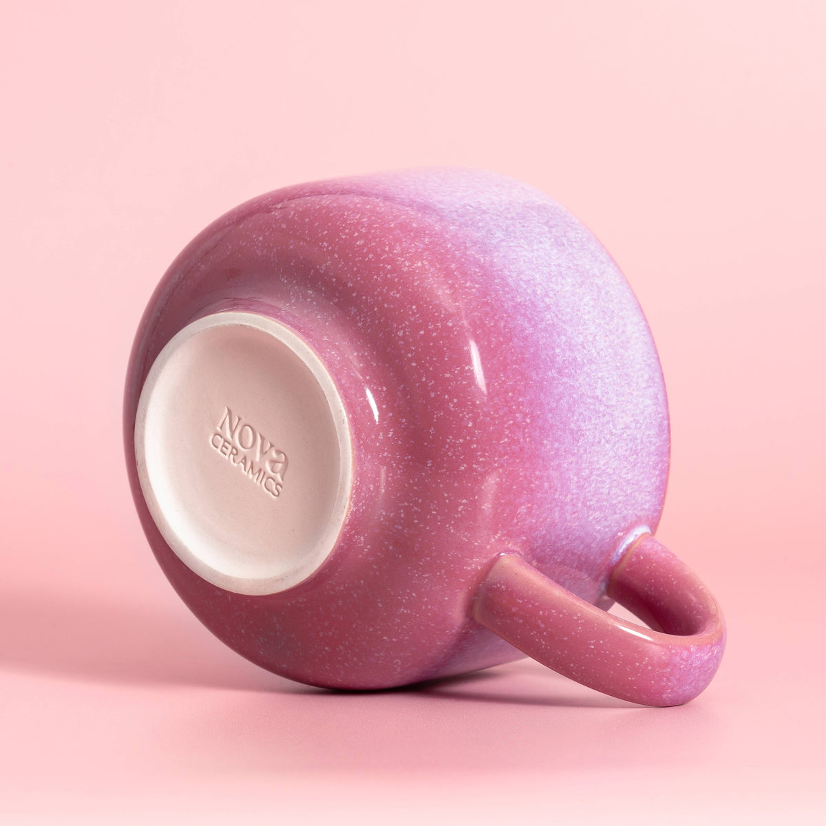 A small ceramic mug with a pink and purple glittery gradient glaze, shown from a side angle on a solid pink background. The inside of the mug features a white base with the brand name 'NOVA CERAMICS' embossed.