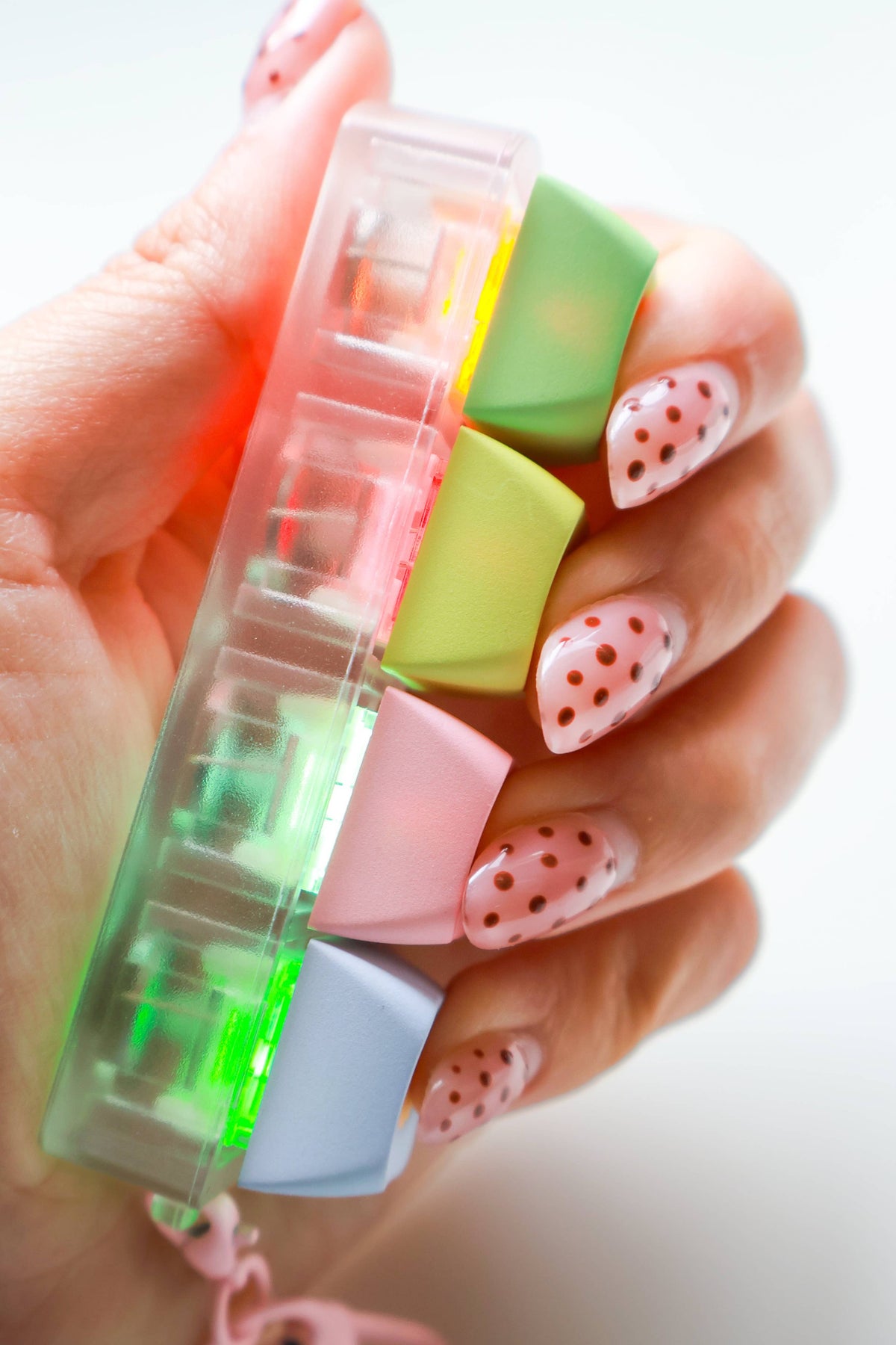 A hand with pink strawberry-patterned nails holds a translucent plastic organizer containing pastel-colored square inserts in green, pink, yellow, and light blue against a bright white background.