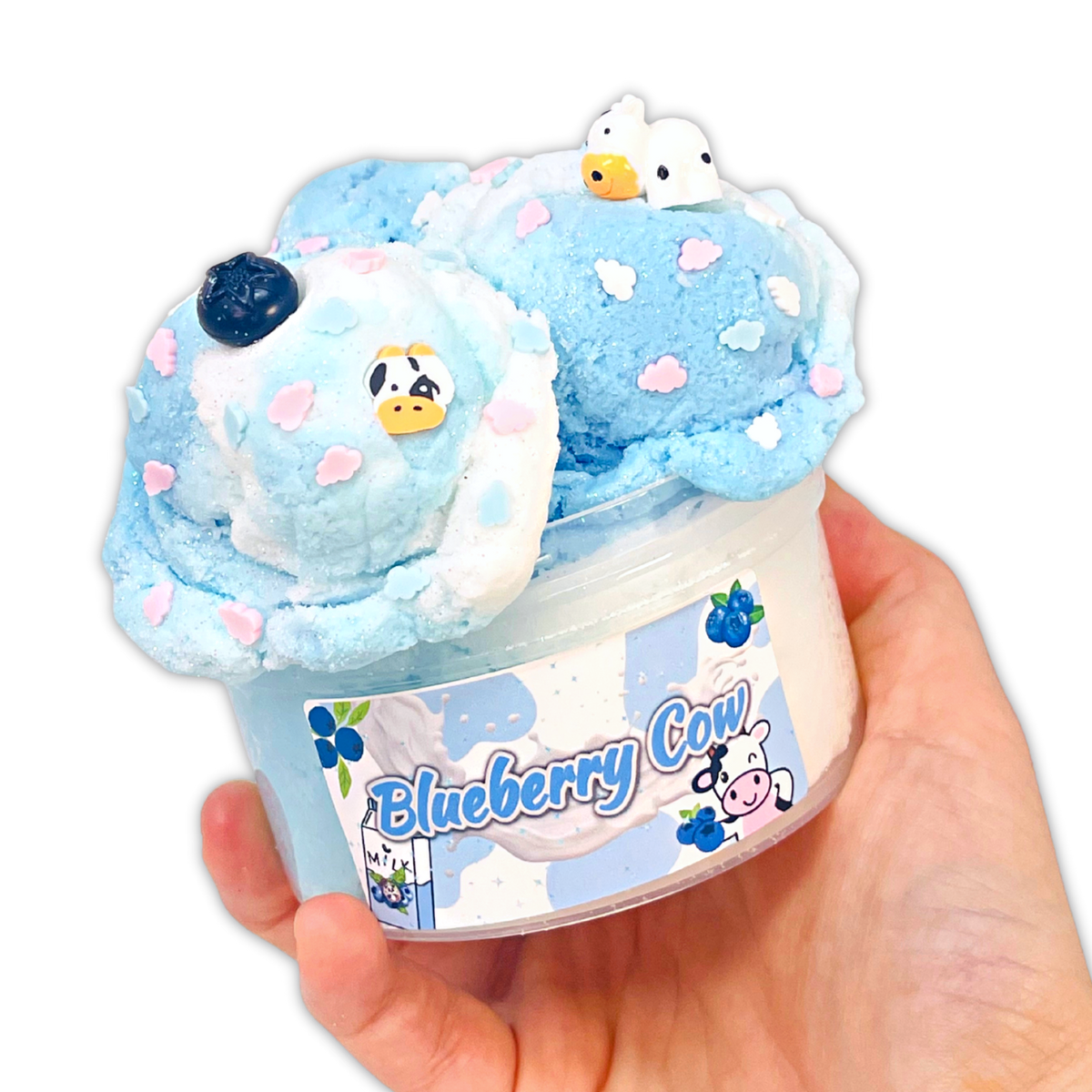 A hand holding a clear plastic tub filled with light blue and white whipped cream or frosting topped with decorative blueberry and cow motifs, with a label reading 'Blueberry Cow' on a plain white background.
