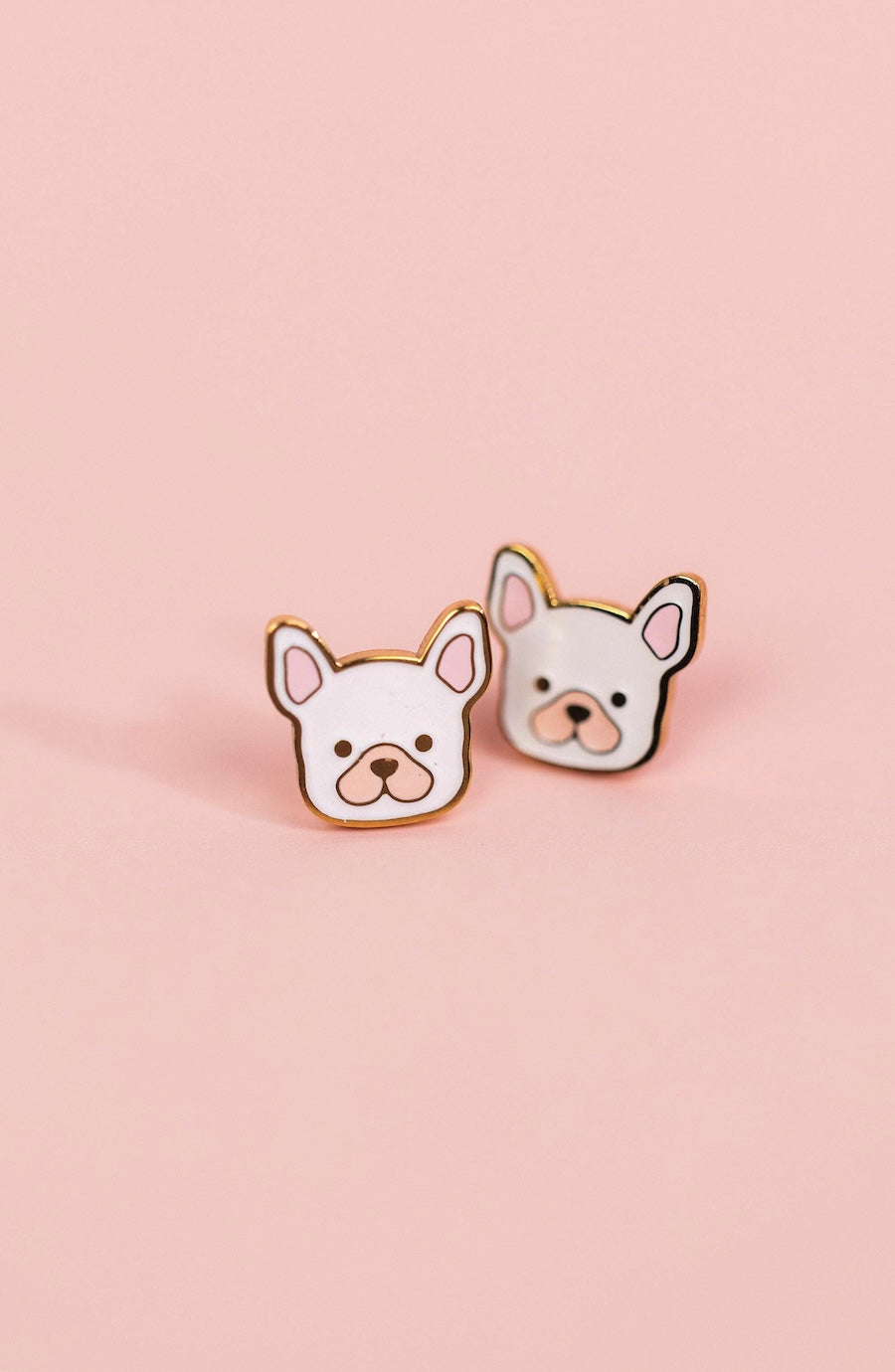 A pair of stud earrings shaped like cute white French Bulldog faces with gold-tone outlines, set against a solid pale pink background.