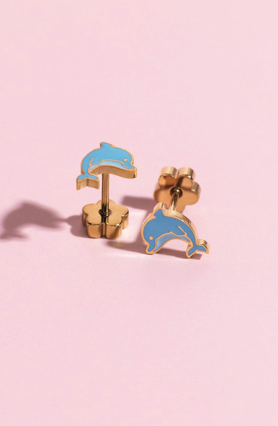 A pair of gold-tone dolphin stud earrings with light blue enamel fills, displayed on a soft pink background.