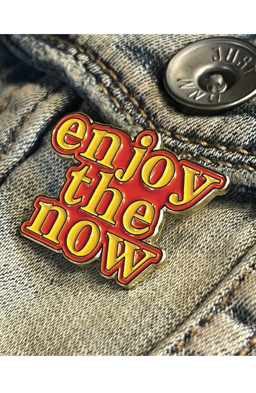 Enamel pin with 'enjoy the now' text on denim fabric