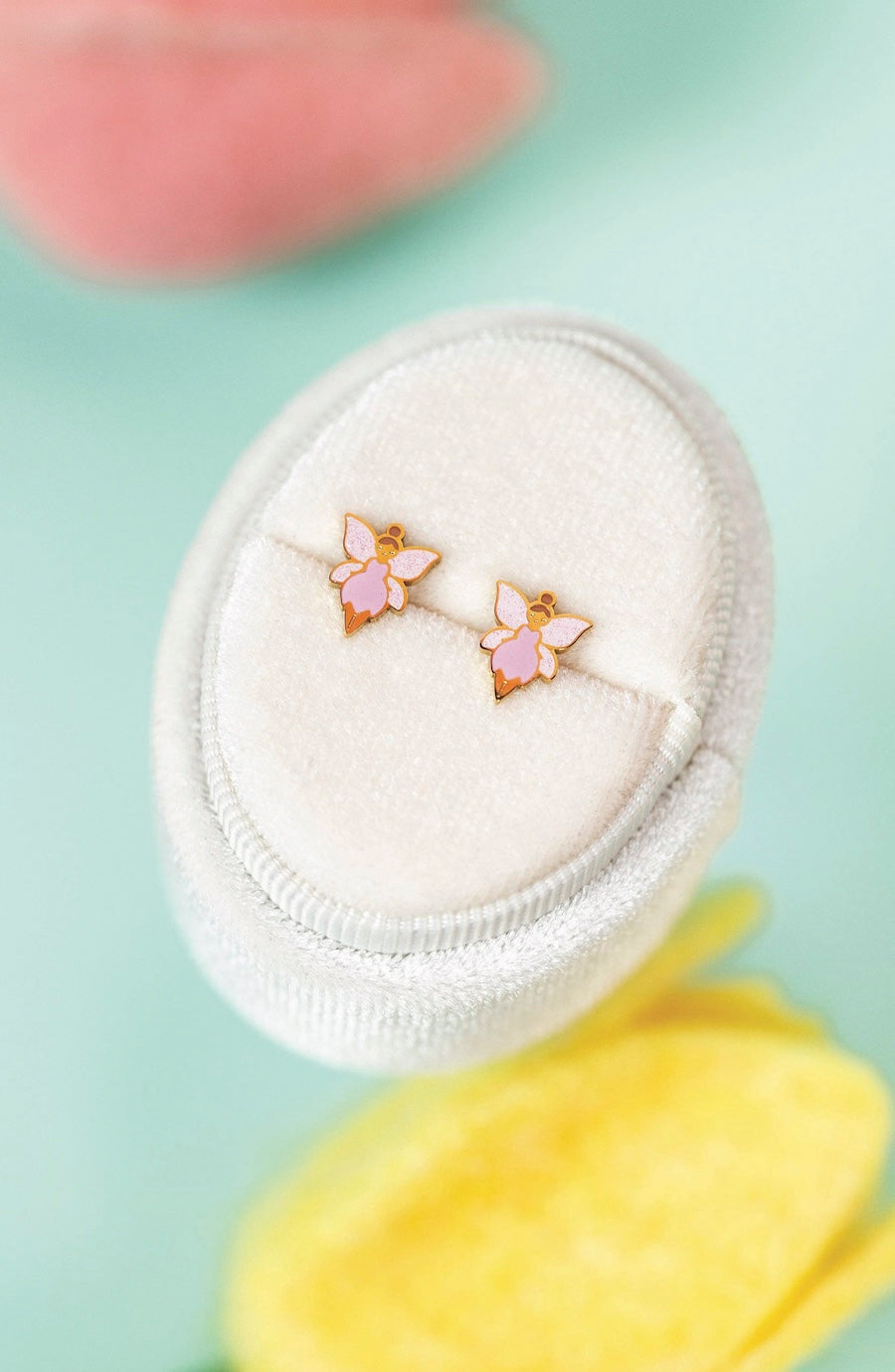 A pair of small butterfly stud earrings with pink enamel wings and gold-toned metal outlines, displayed on a white oval jewelry stand against a soft mint background with blurred pink and yellow floral shapes.