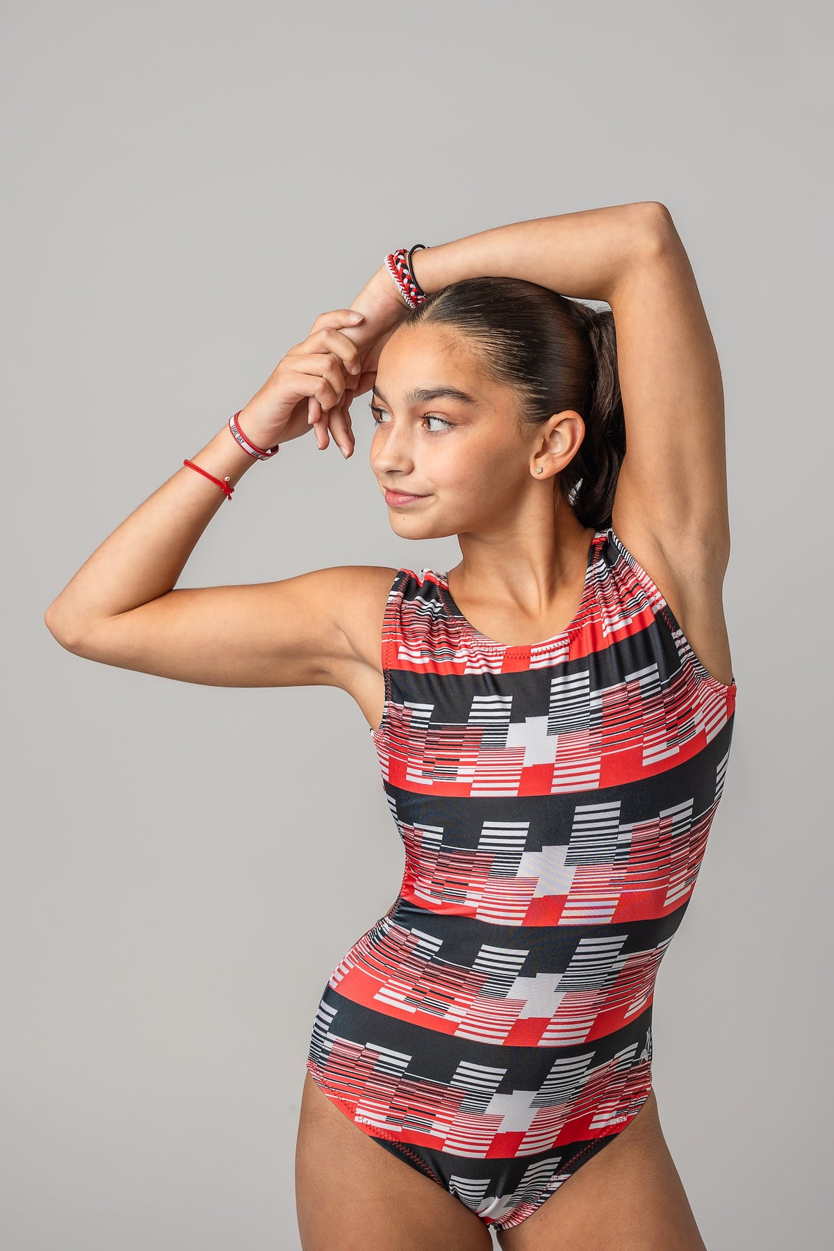 A young female gymnast modeling a sleeveless leotard for girls with a red, black, and white geometric pattern, posing against a solid gray studio background.
