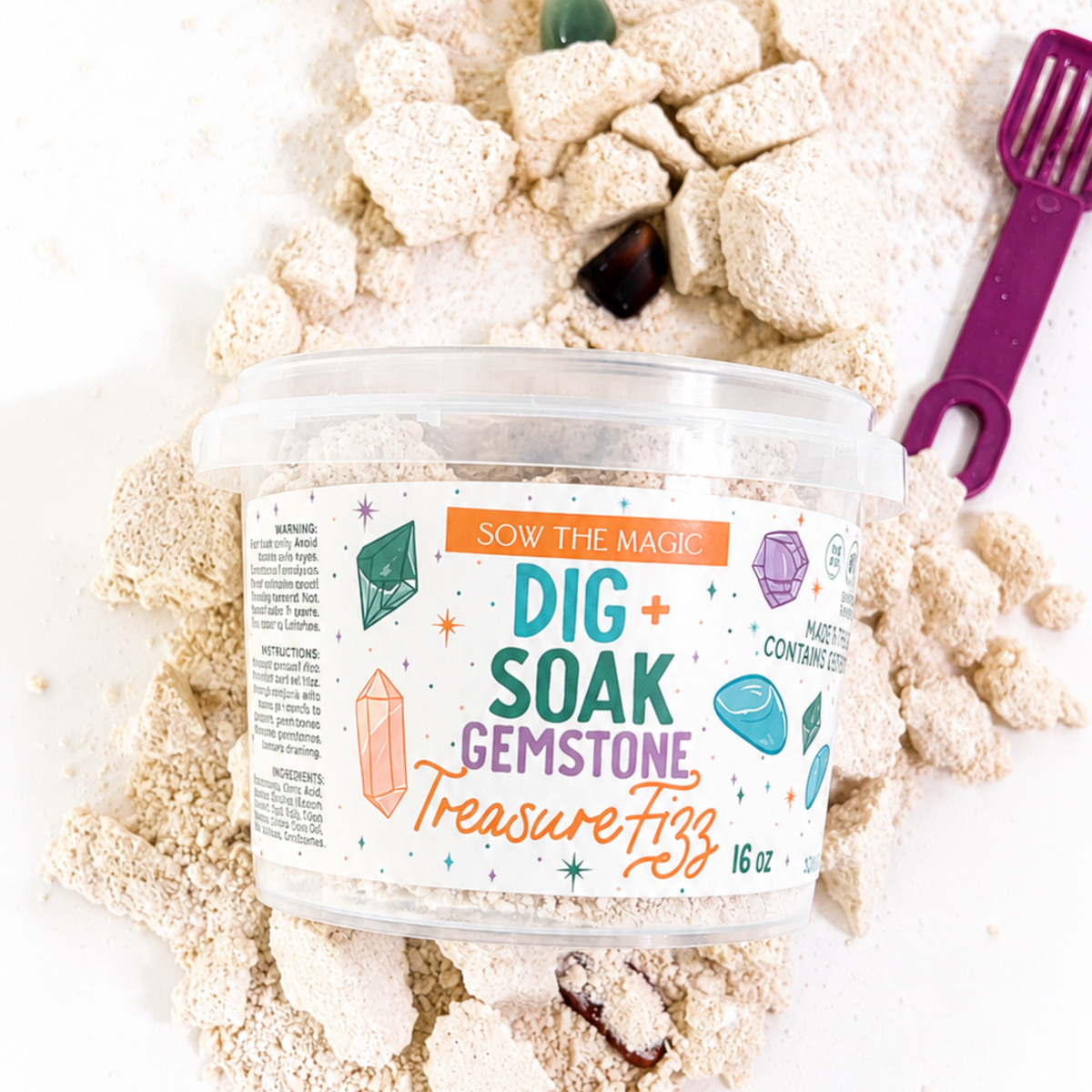 Container labeled 'Dig Soak Gemstone Treasure Fizz' surrounded by sand and small stones on a white background.