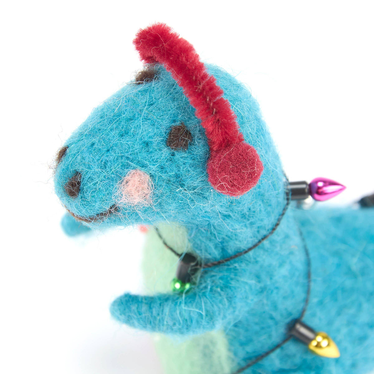 A close-up of a blue felted wool dinosaur plush toy wearing red fuzzy earmuffs and a string of holiday lights, set against a clean white background.