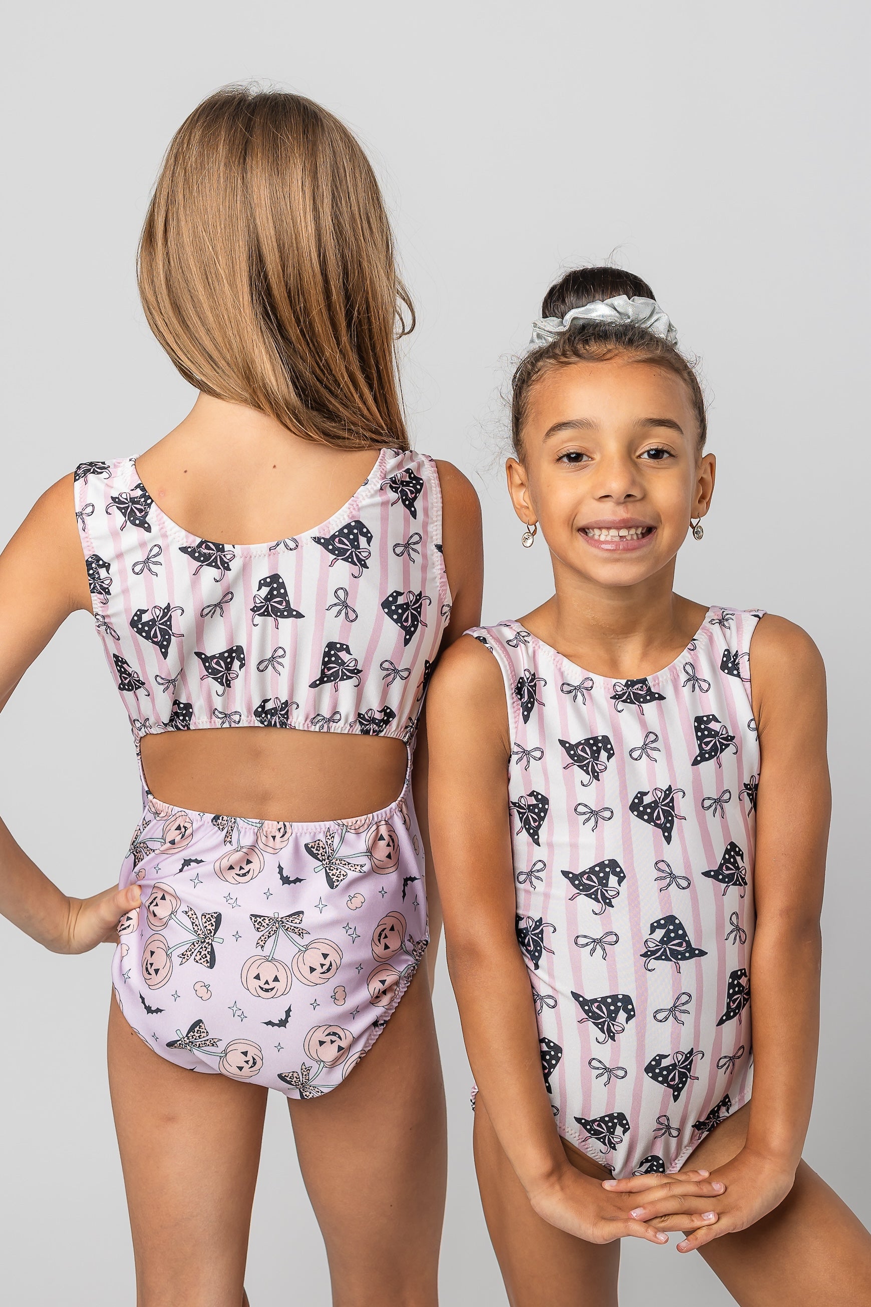 Two young girls modeling matching sleeveless leotards, shown from the back and front against a light gray studio background. The leotards have a pink and white striped base with black witch hat illustrations and small butterfly motifs.