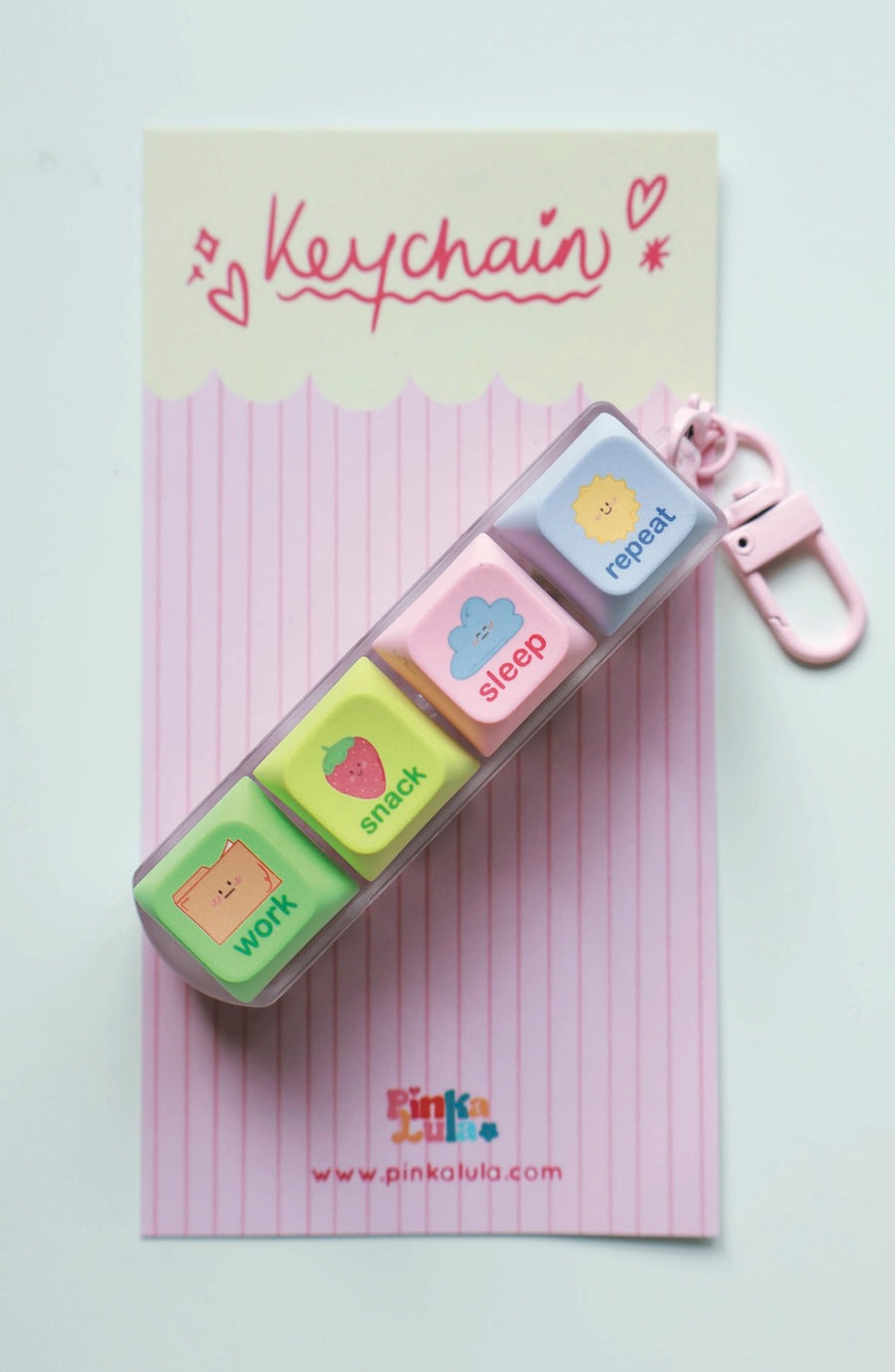 A pastel-colored keychain container with four labeled cubes (work, snack, sleep, repeat) resting on its pink striped backing card, which has 'Keychain' written on it.