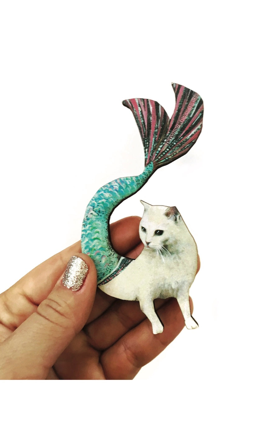 A hand holding a decorative metal pin shaped like a white cat sitting on a green and pink mermaid tail, against a plain white background.