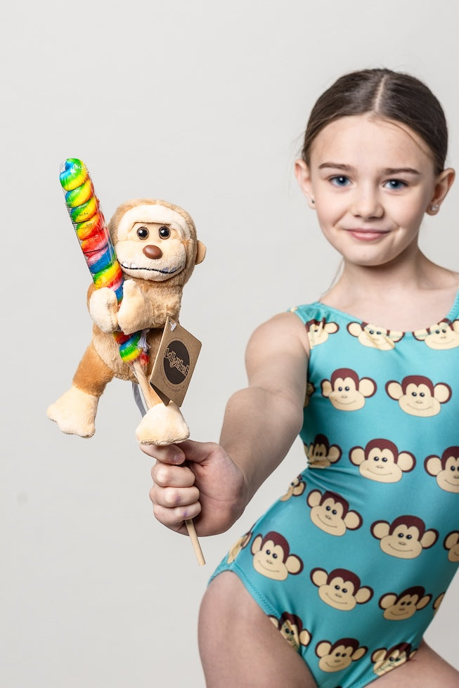 Young girl in a monkey-patterned leotard holding a colorful monkey toy on a rainbow sucker against a plain background