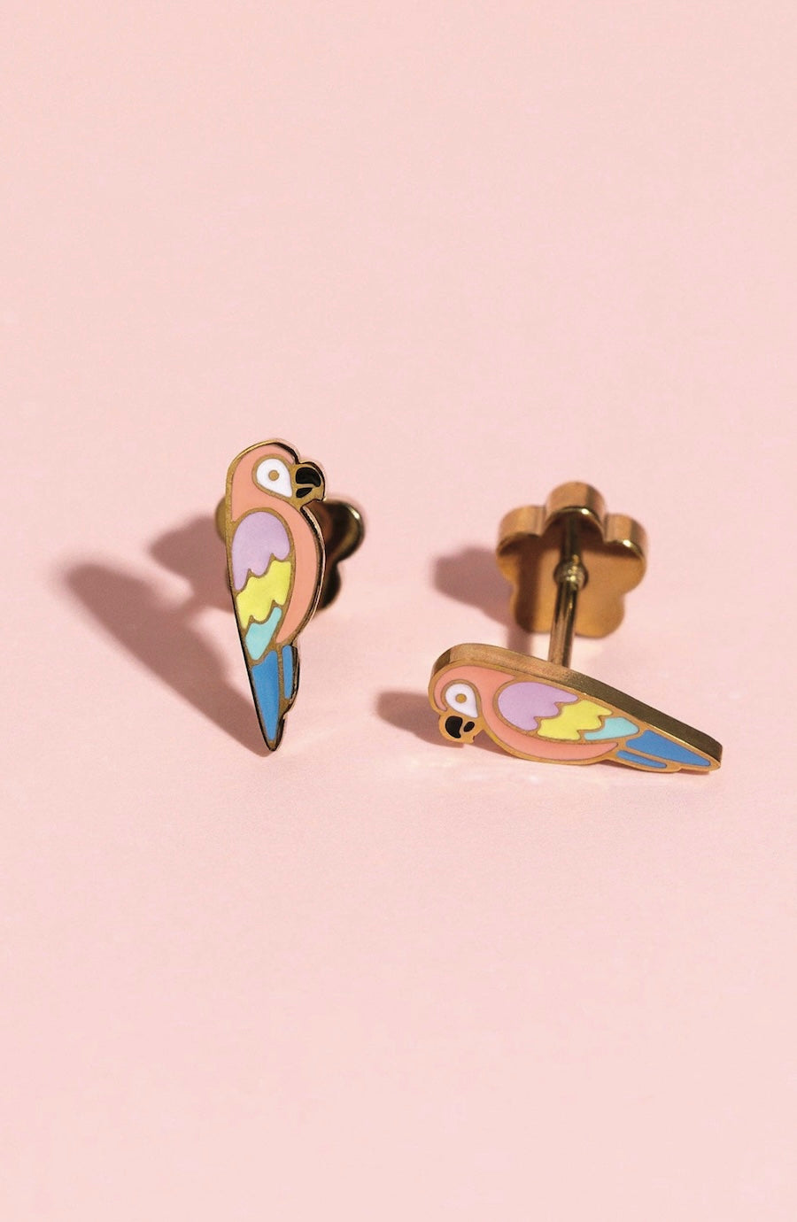 A pair of gold-toned stud earrings shaped like colorful parrots, featuring pastel yellow, purple, blue, and green enamel details, displayed on a soft pink background.