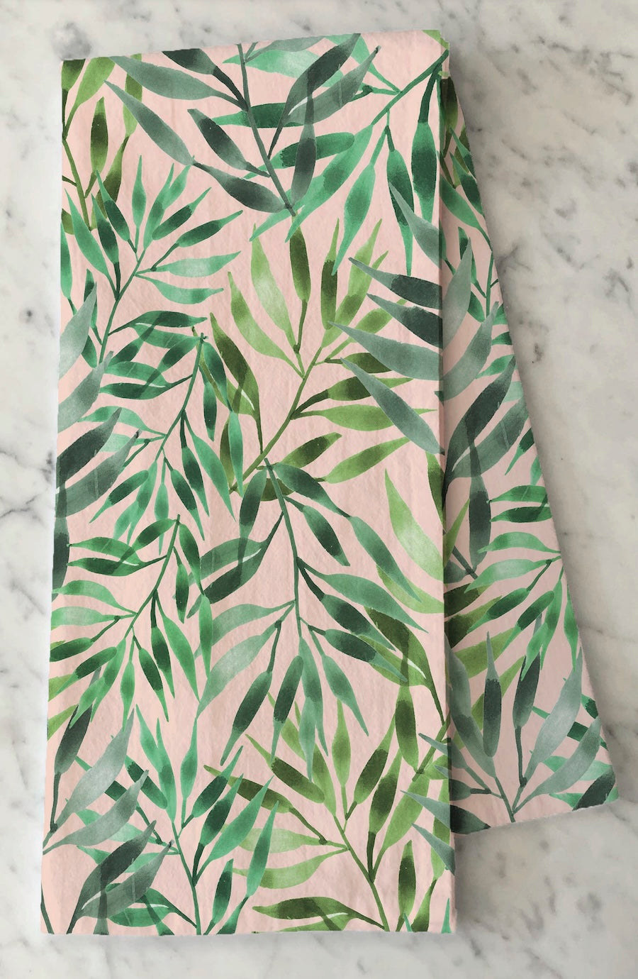 A folded fabric item, likely a napkin or table runner, featuring a watercolor-style green palm leaf pattern on a soft blush pink background, displayed on a white marble surface.