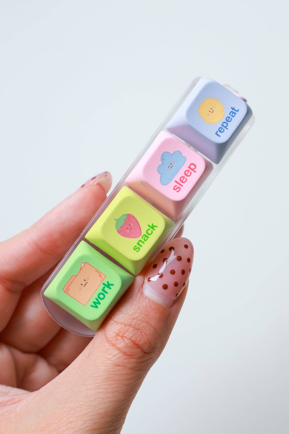 A hand holding a clear plastic case containing four pastel-colored square keys labeled repeat, sleep, snack, and work, each with a small cartoon face.