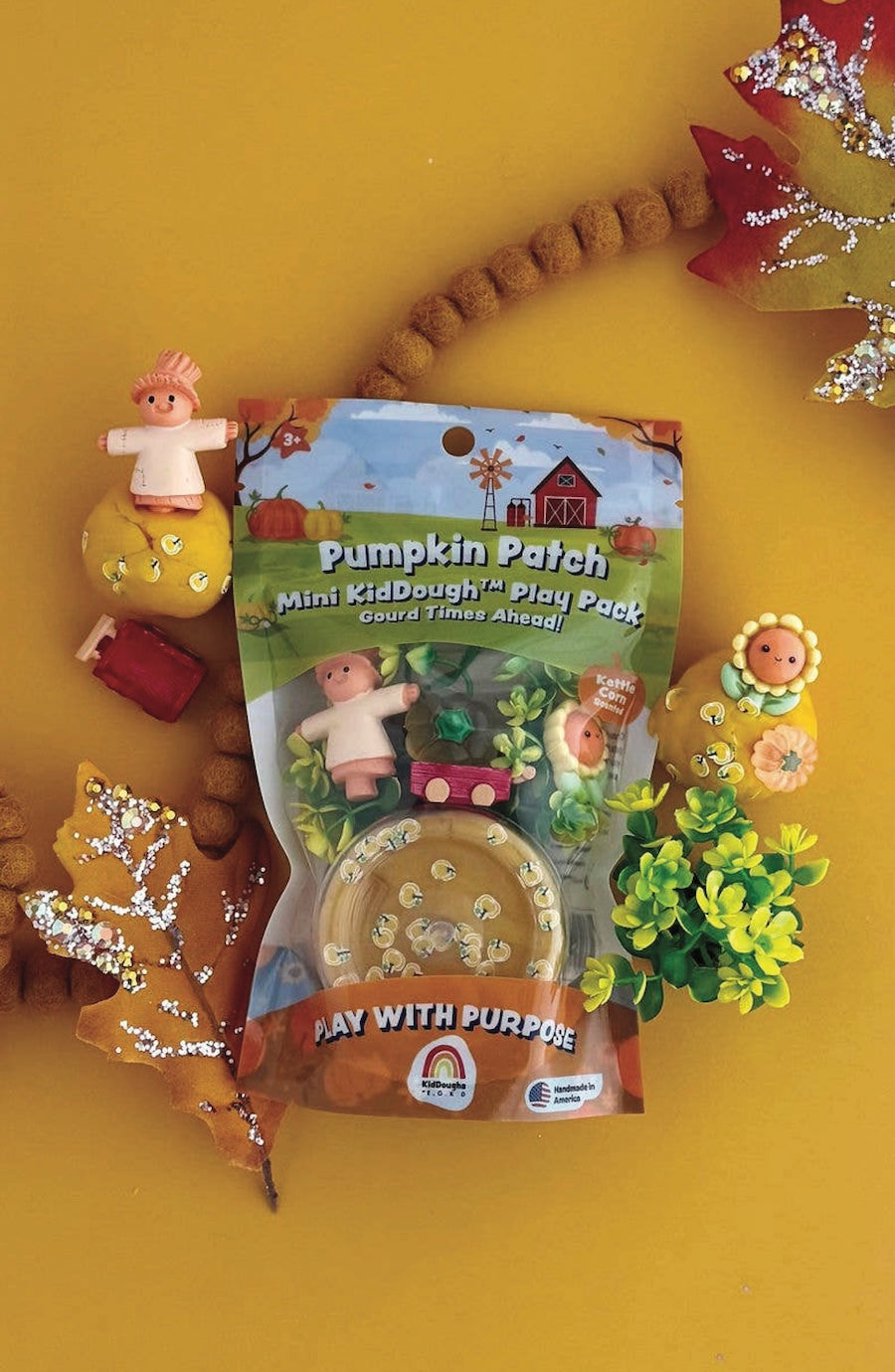 Pumpkin Patch Mini KidDough Play Pack toy set in its packaging, surrounded by autumn-themed decorative props on a solid yellow background.