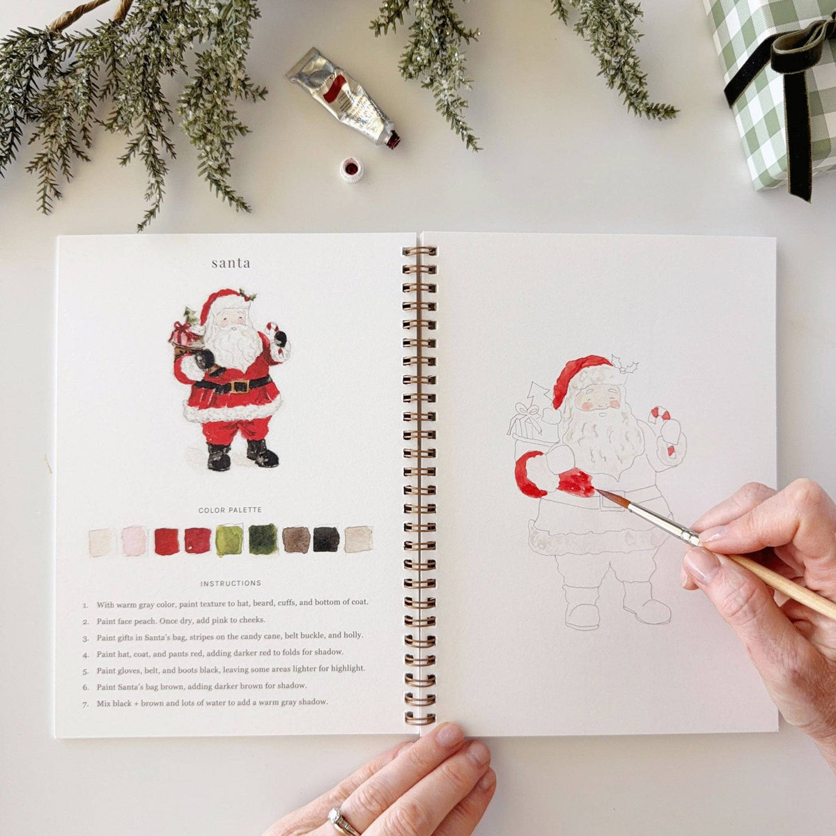 An open spiral-bound coloring book or art journal laid on a white surface, showing a Santa illustration with color palette and instructions on the left page, and a partially painted outline of Santa on the right page. A hand holds a paintbrush over the right page, and holiday greenery, a tube of paint, and a checkered fabric corner are visible around it.