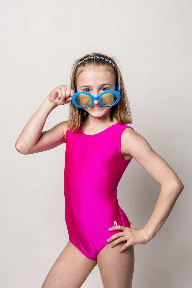Young dancer in a solid magenta pink leotard wearing blue goggles against a plain background