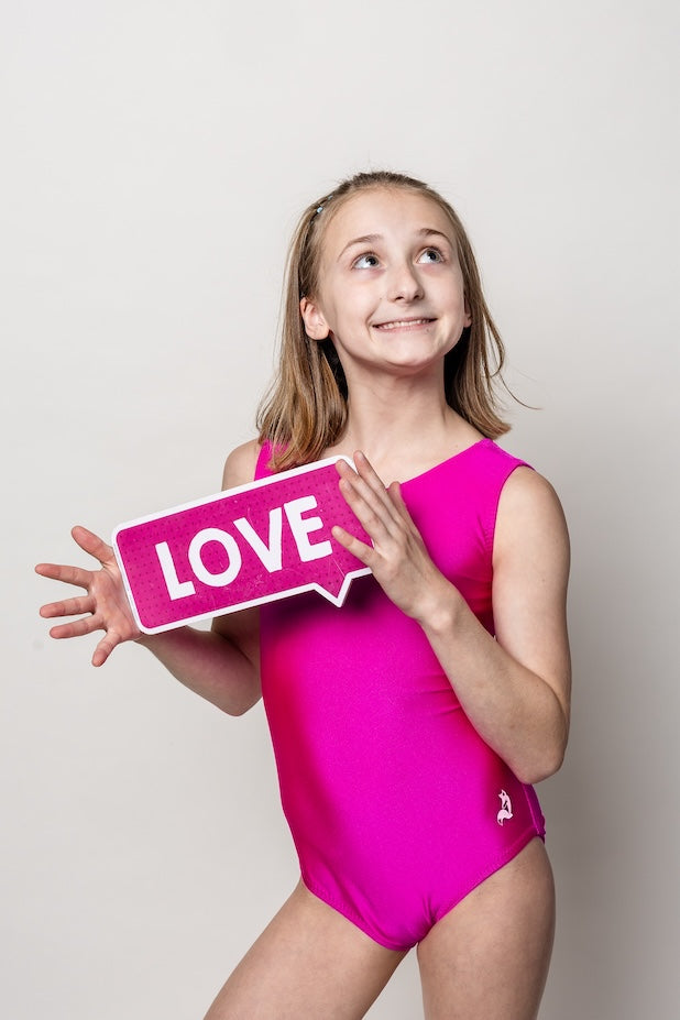 Young gymnast in a solid pink magenta gymnastics leotard holding a 'LOVE' sign against a plain background