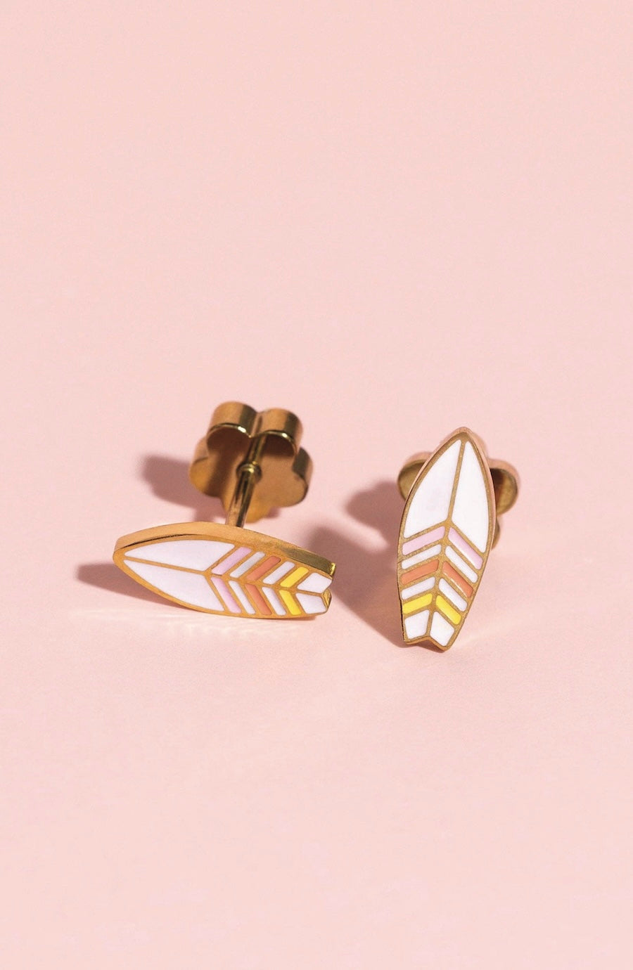 A pair of gold-toned stud earrings with white enamel inlays and chevron patterns in gold and light pink, displayed on a soft pink background.