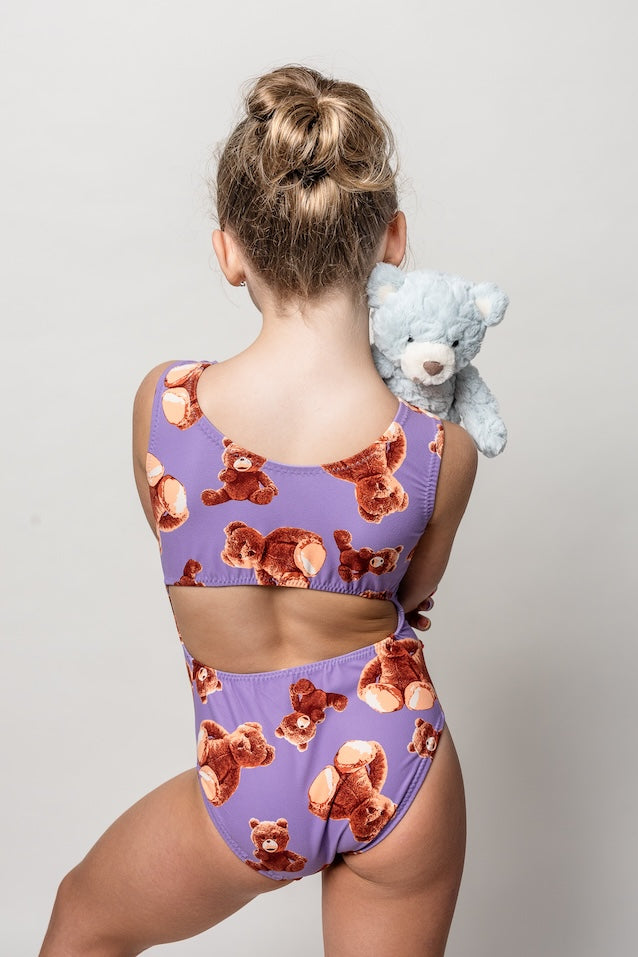 young gymnast wearing a purple teddy bear patterned gymnastics leotard holding a baby blue teddy bear against a plain background