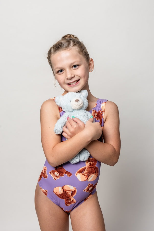 Young gymnast in a ultra soft purple leotard with plush teddy bear pattern holding a stuffed teddy bear against a plain background