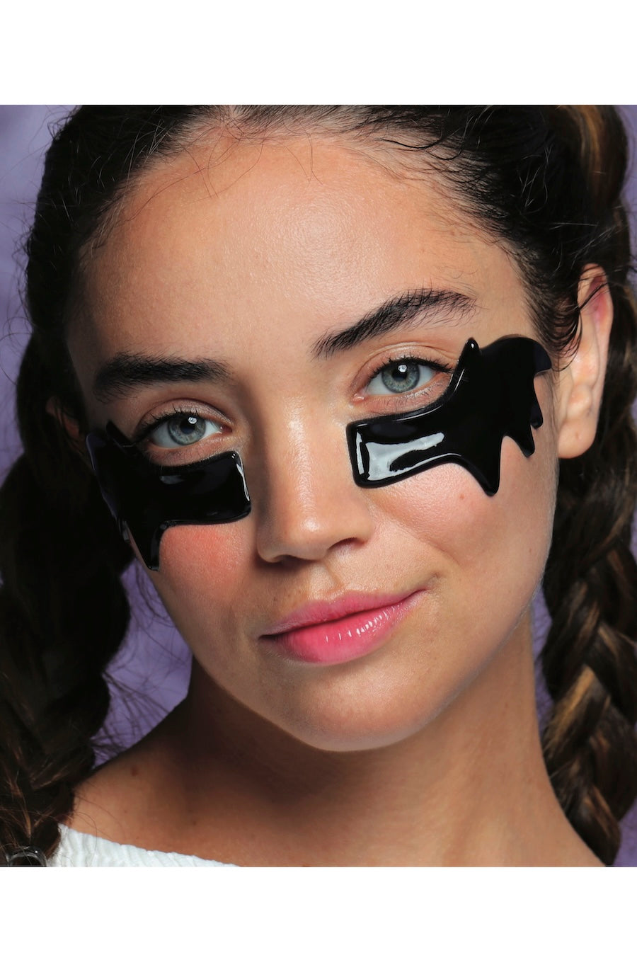 A close-up of a young woman with black eye patches applied around her eyes, shown against a purple background.