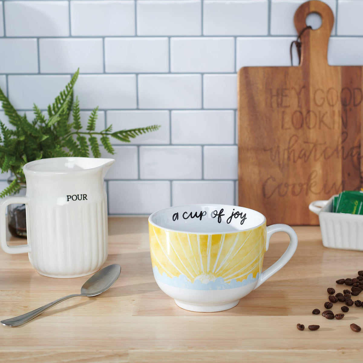 Mug with 'a cup of joy' text on a kitchen counter with a pitcher, spoon, and coffee beans.